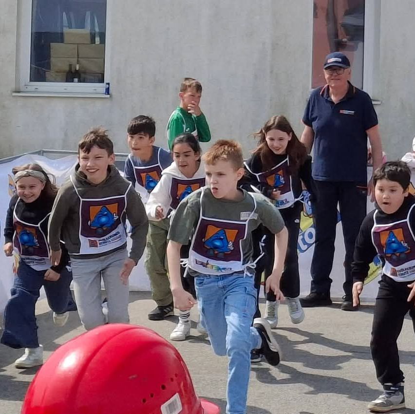 Eine Gruppe von Kindern in Schürzen und Turnschuhen rennt auf einer Straße. Ein Mann in einem blauen Shirt und einem Hut beobachtet sie. Ein roter Helm ist im Vordergrund.