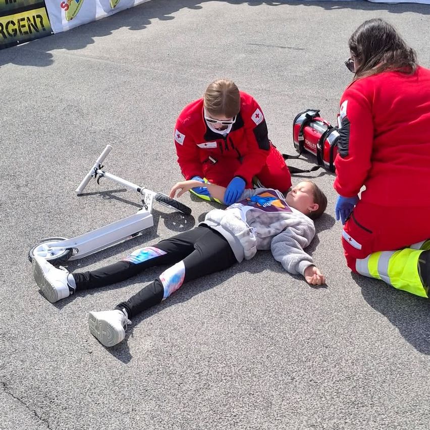 Ein junges Mädchen liegt auf dem Boden mit einem Roller in der Nähe. Zwei Personen in roten Uniformen kümmern sich um sie, eine kniet und die andere steht. Eine rote medizinische Tasche ist neben ihnen.