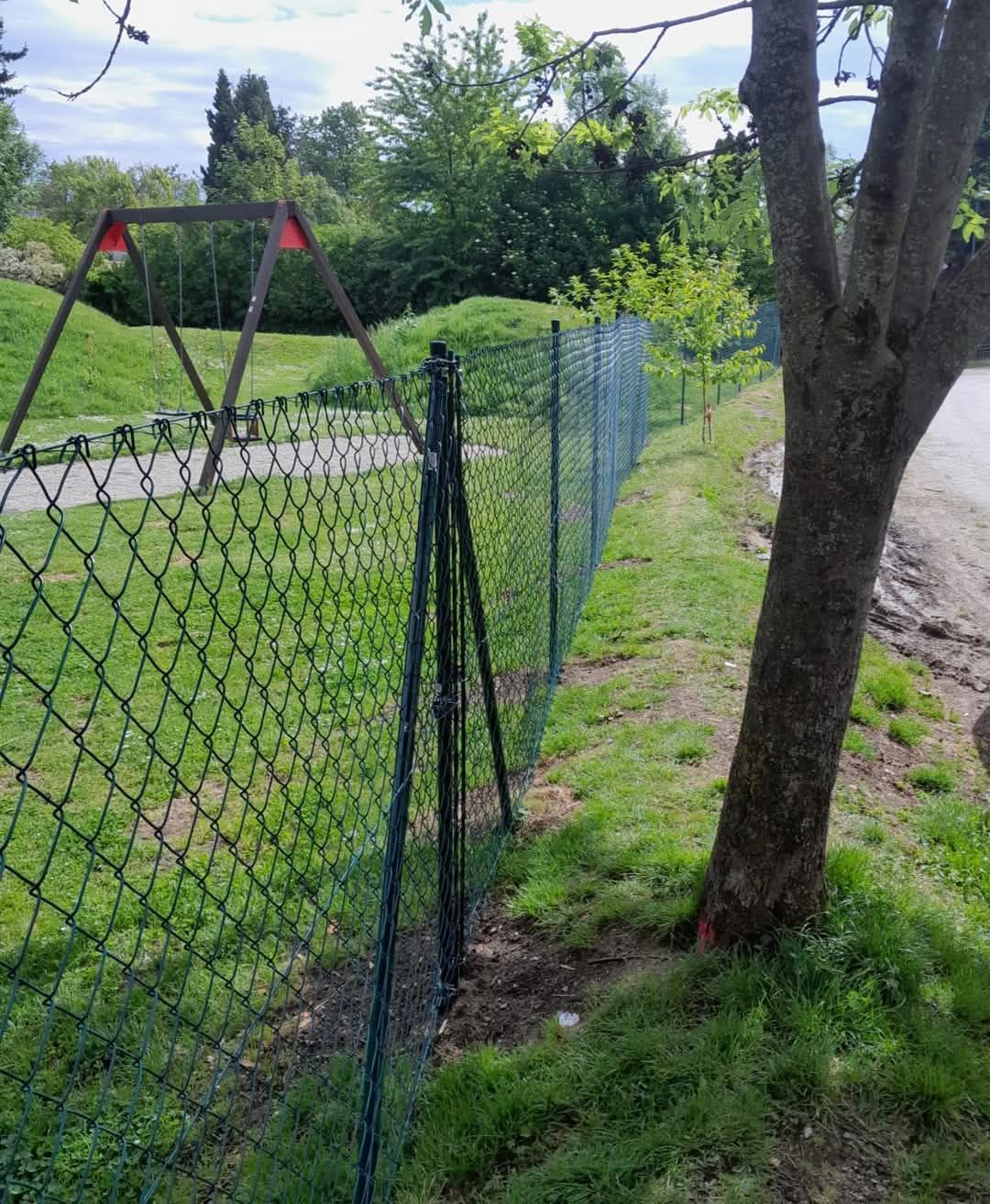 Ein grüner Maschendrahtzaun grenzt an einen Rasenbereich mit einem Baum und einer Holzschaukel in der Ferne.
