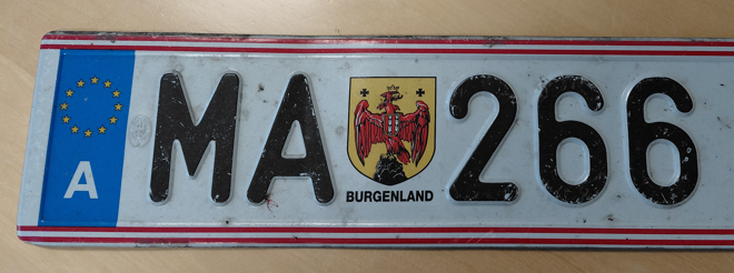 Ein weißer Nummernschild mit rotem Rand, einem blauen Streifen auf der linken Seite und einem gelben Wappen in der Mitte. Das Schild hat die Buchstaben MA und die Zahlen 266.