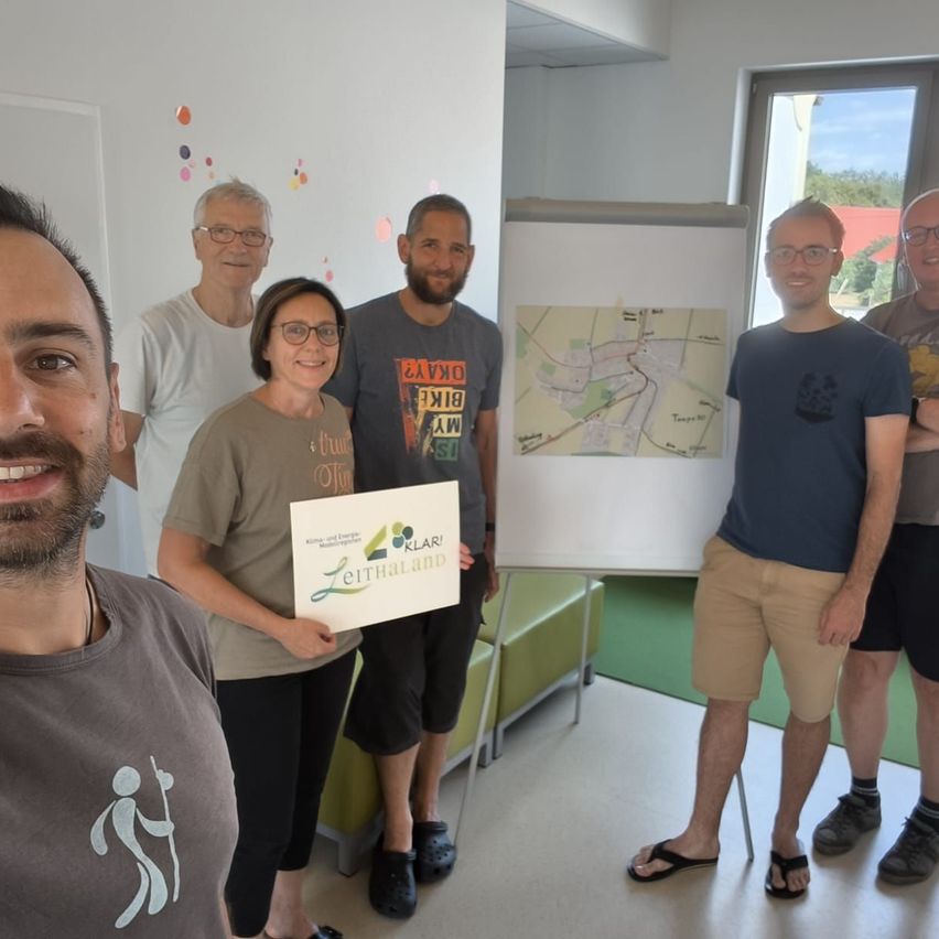 Eine Gruppe von Menschen steht in einem Raum mit einer Karte auf einer Tafel. Eine Frau hält ein Schild mit dem Text 'Leitland'.