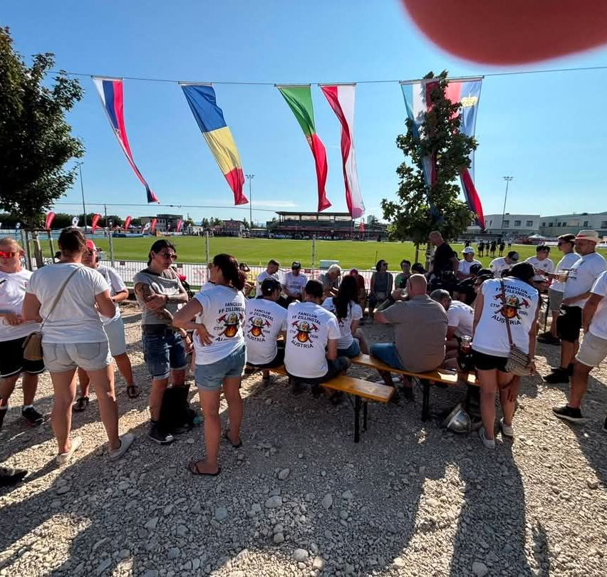 Eine Gruppe von Menschen in weißen T-Shirts mit Text und Logos steht und sitzt um einen Tisch in einem Außenbereich mit Flaggen und einem Baum.
