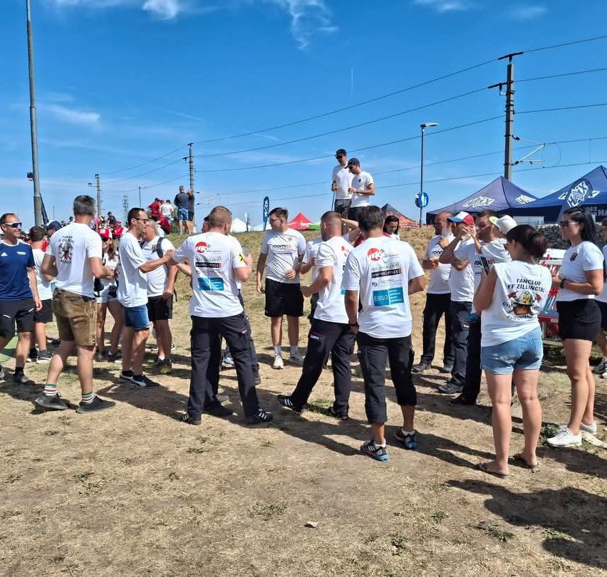 Eine Gruppe von Menschen in passenden weißen T-Shirts versammelt sich auf einem Erdfeld, einige stehend und andere gehend, mit einem klaren blauen Himmel im Hintergrund.