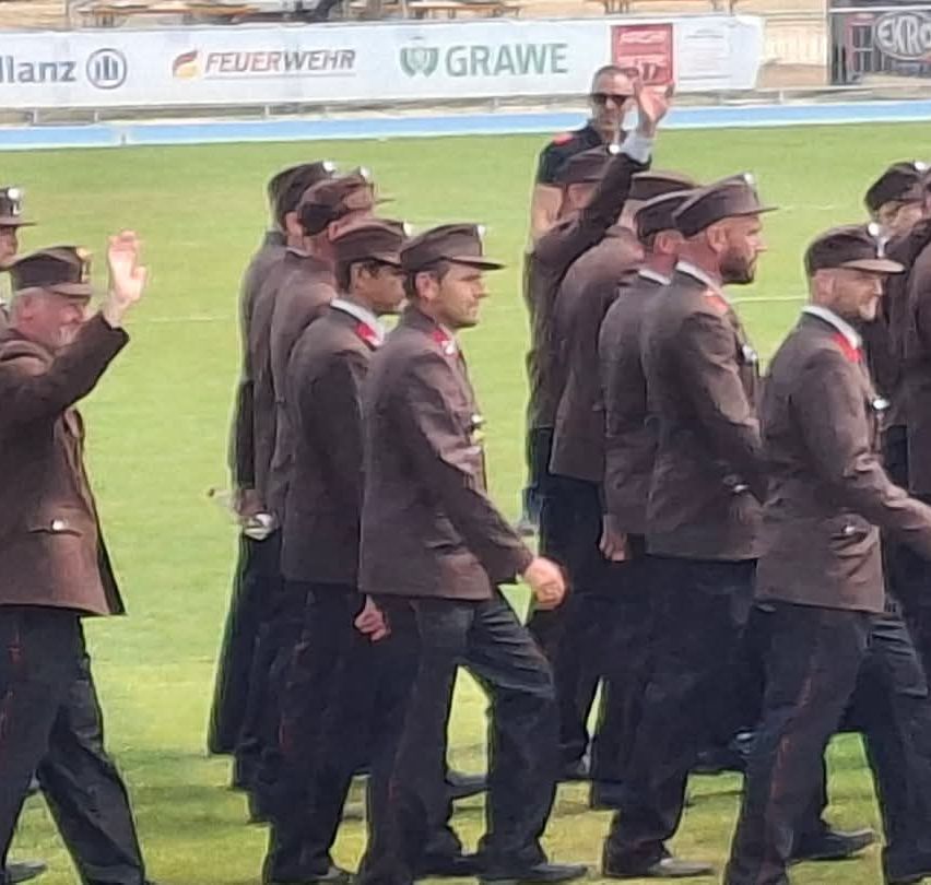 Eine Gruppe von Männern in Uniform, die auf einem grünen Feld gehen, einige winken, andere schauen nach vorne, mit einem verschwommenen Hintergrund von einem Zaun und Werbetafeln.
