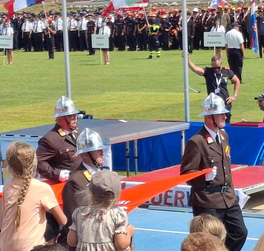 Eine Gruppe von Feuerwehrleuten in Uniform steht auf einer Bühne mit Flaggen dahinter. Einige halten Flaggen, andere stehen in Formation. Eine Menschenmenge beobachtet.