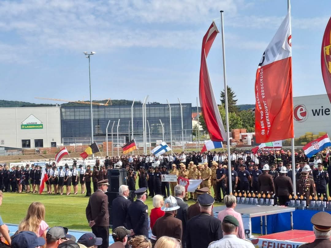Eine große Versammlung von Menschen in Uniform, möglicherweise eine Parade, mit verschiedenen Flaggen und einer Bühne im Vordergrund.