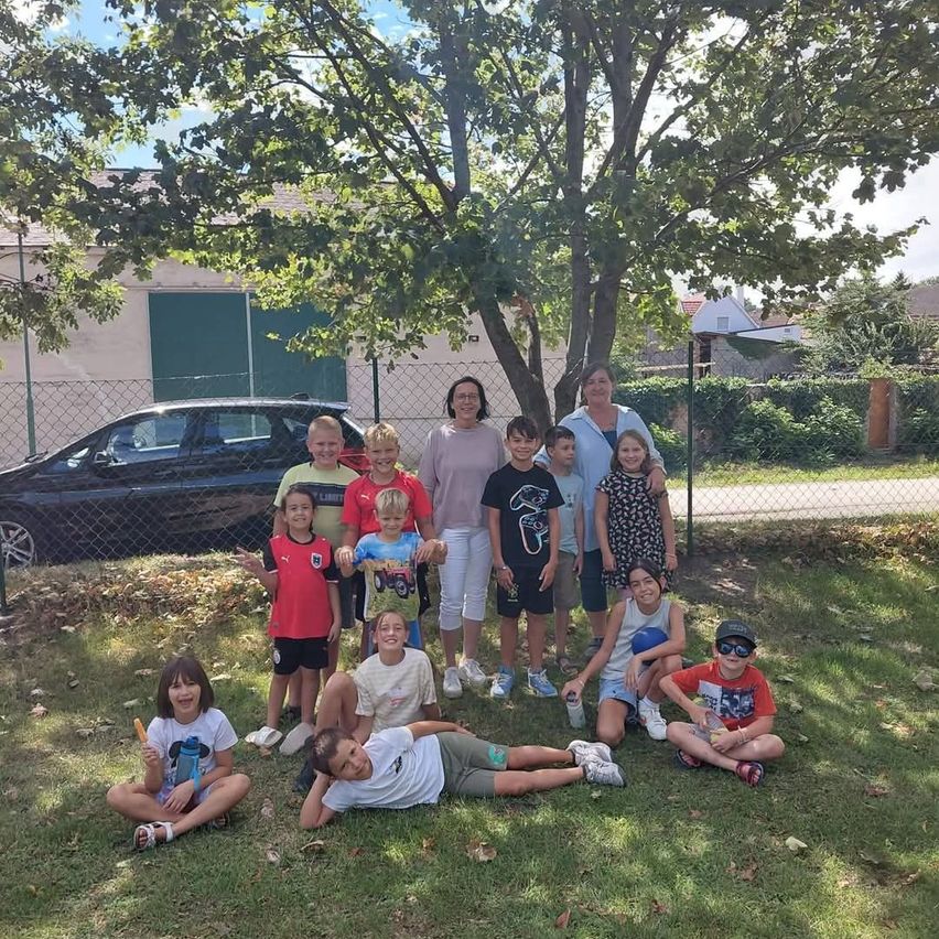 A group of children and two adults pose for a photo in a park, with a tree and a car in the background.