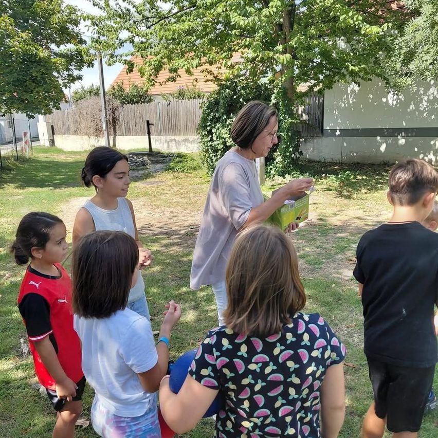 A woman with glasses holds a green box and speaks to a group of children in a yard. Trees and a house are in the background.