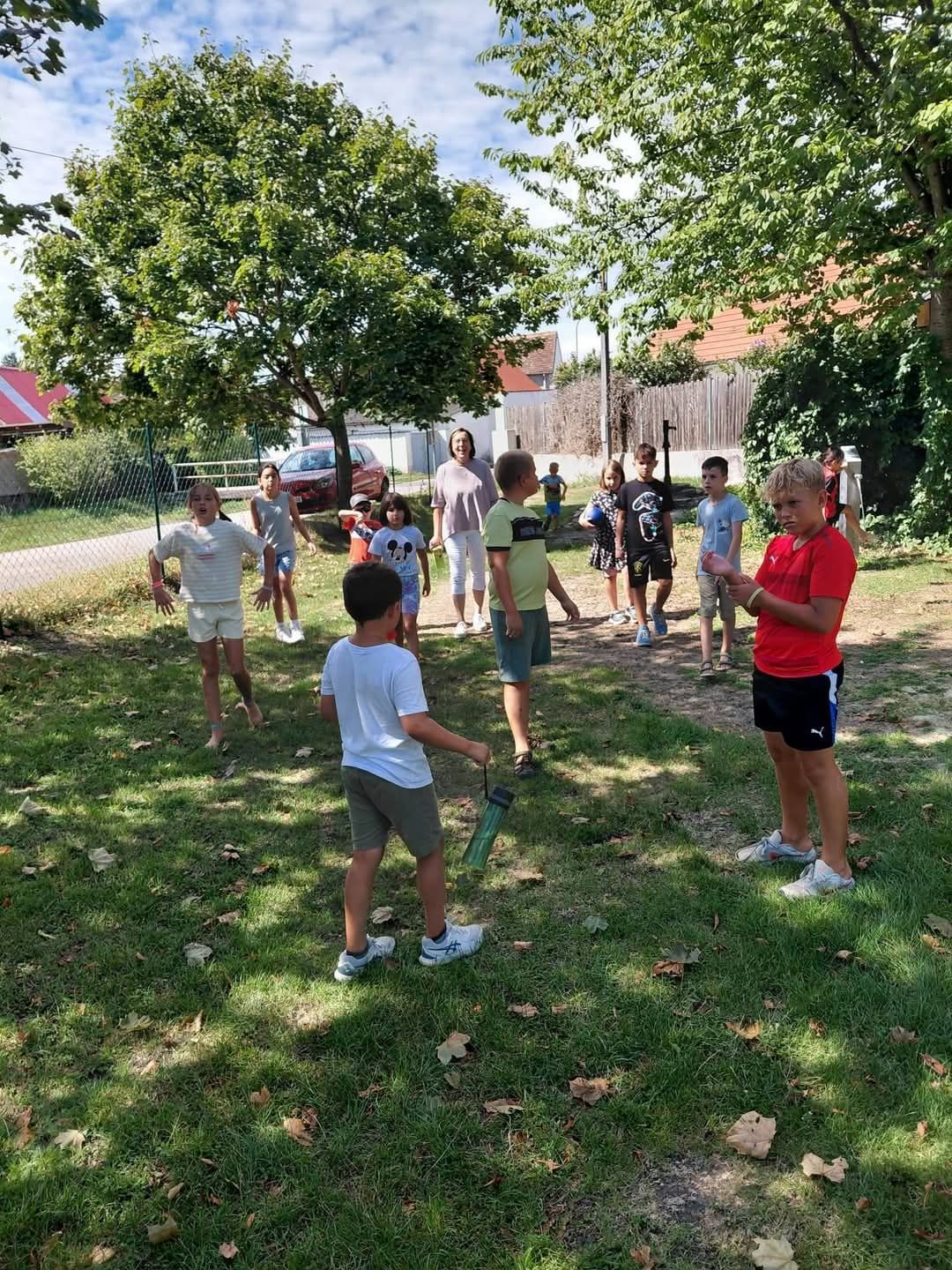 A group of children and adults play on a grassy field, some holding water bottles, with trees and a fence in the background.