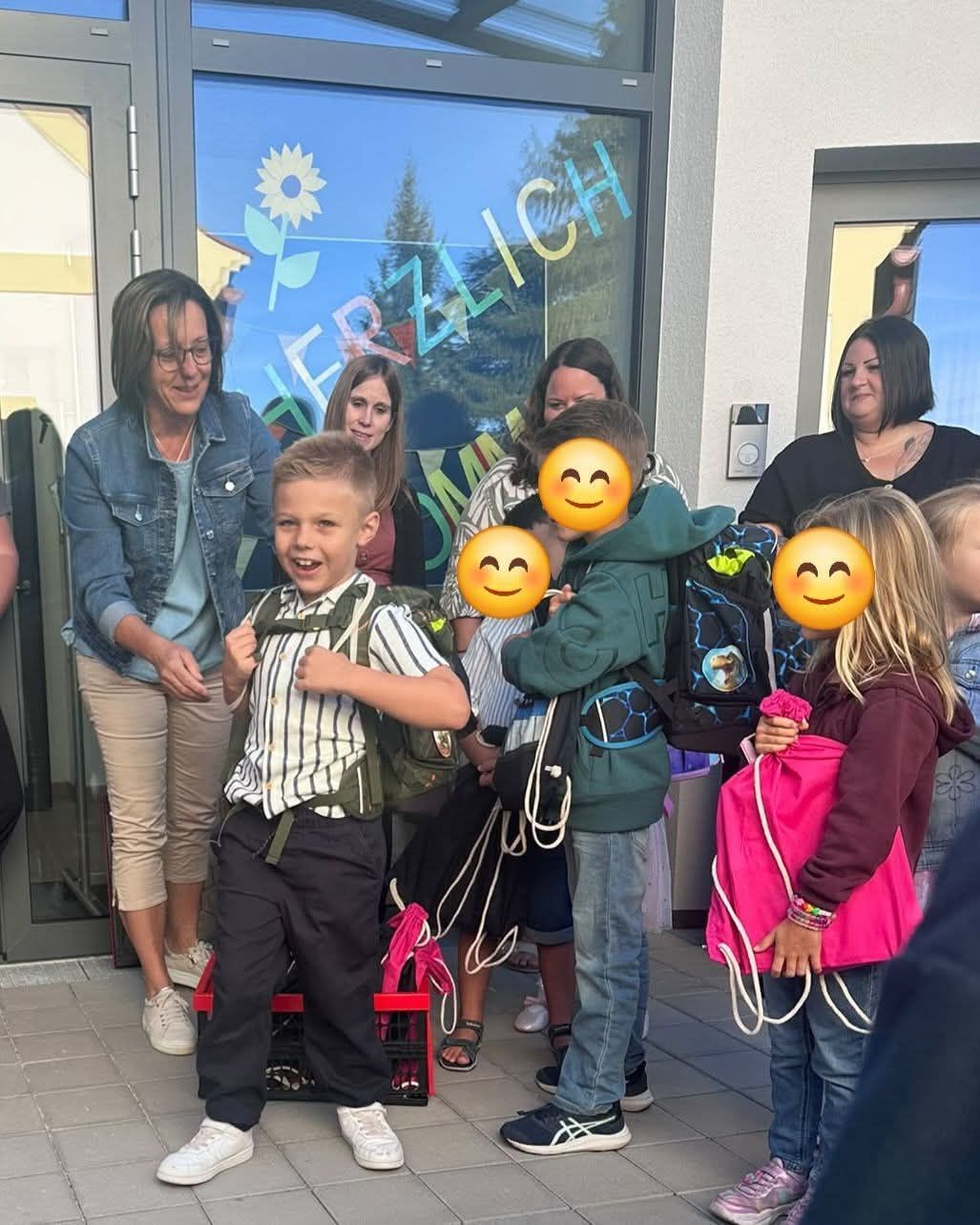 A group of children and adults stand outside a building, some with backpacks and smiling faces. A woman in a denim jacket stands behind a boy with a striped shirt.