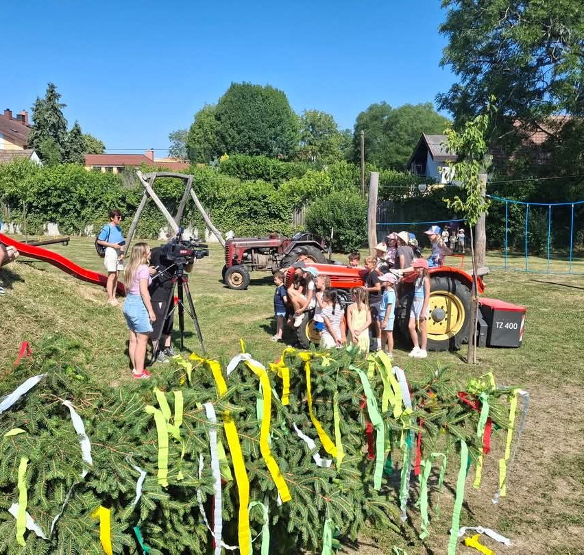 Eine Gruppe von Kindern versammelt sich an einem sonnigen Tag um einen roten Traktor, während eine Person filmt. Im Hintergrund befinden sich Bäume und ein Haus.