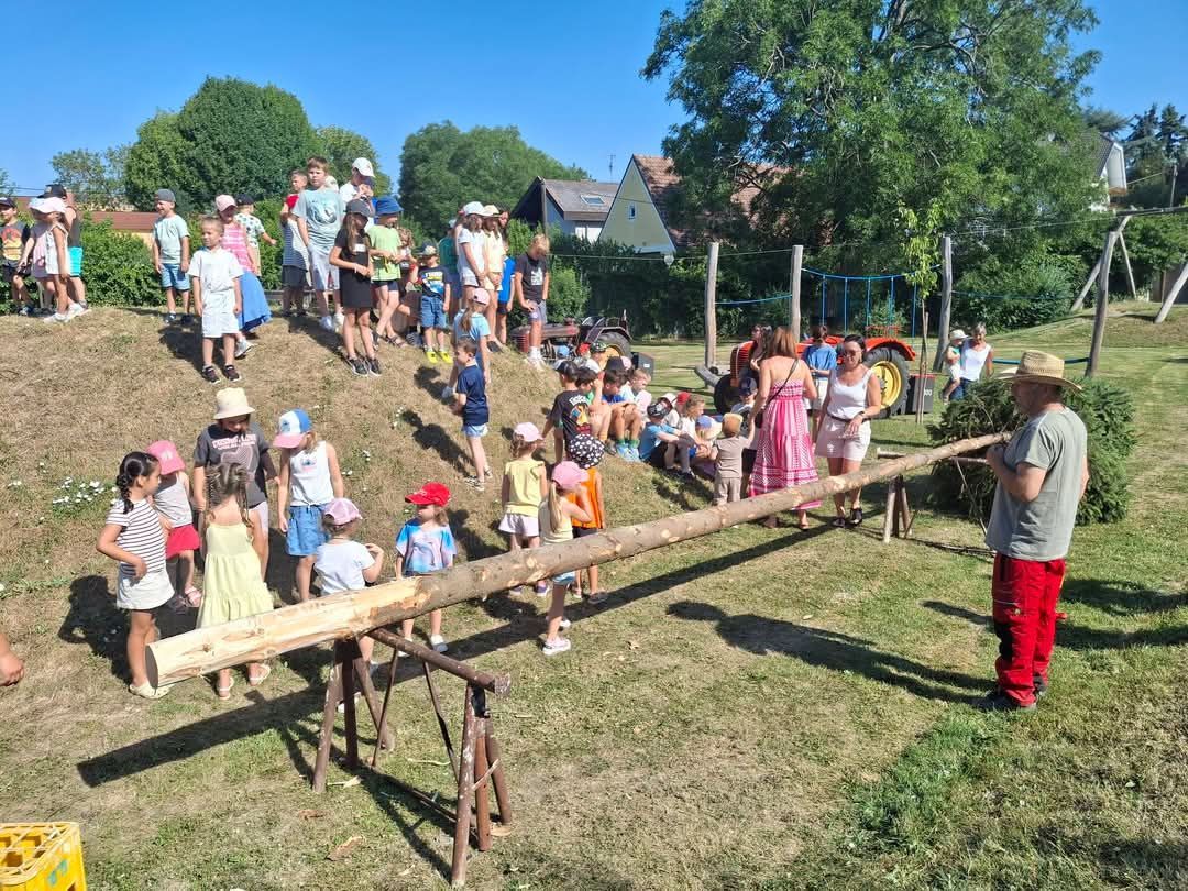 Eine Gruppe von Kindern und Erwachsenen ist im Freien. Sie stehen und sitzen auf einem schiefen Boden mit einem Baumstamm. Sie scheinen eine Aktivität zu machen.
