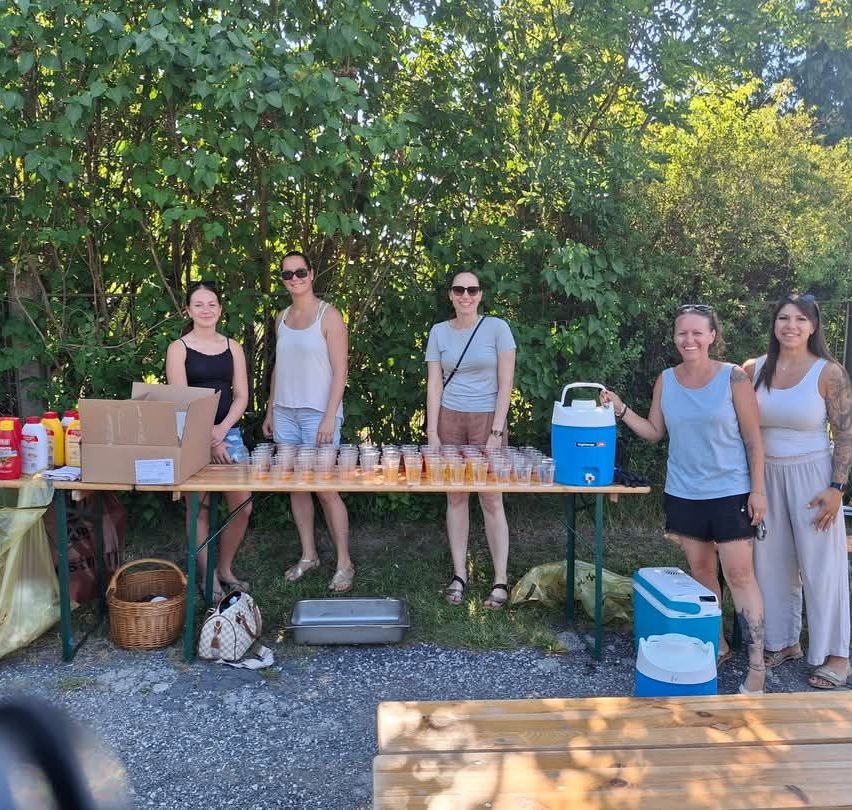 Fünf Frauen stehen bei einem Tisch mit Getränken, einem Kühler und Kartons. Sie befinden sich in einem Außenbereich mit Bäumen im Hintergrund.