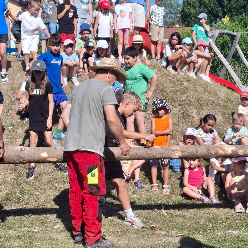 Mehrere Kinder und Erwachsene beobachten einen Mann, der einen Baumstamm sägt. Ein Mann trägt einen Hut und rote Hosen. Ein Kind trägt ein schwarzes T-Shirt.