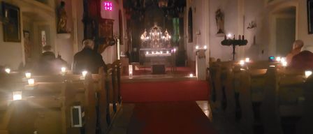 In einer schwach beleuchteten Kirche sitzt ein Mann auf einer Kirchenbank. Der Gang ist mit Teppich ausgelegt und führt zum Altar mit Kerzen. Eine Kandelaber hängt über dem Kopf und eine Statue befindet sich an der Wand.