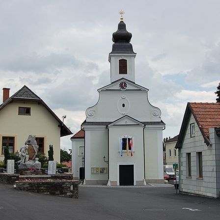 Eine weiße Kirche mit einem schwarzen Turm und einem Kreuz darauf steht vor einem bewölkten Himmel. Flaggen sind am Fenster angebracht.