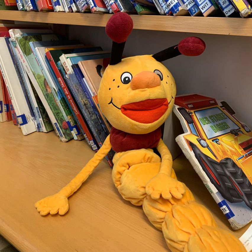 Ein gelbes Plüschtier sitzt auf einem Holzregal umgeben von Kinderbüchern. Das Plüschtier hat große Augen und ein breites Lächeln. In der Nähe befindet sich ein Buch mit einem gelben Baufahrzeug auf dem Cover.