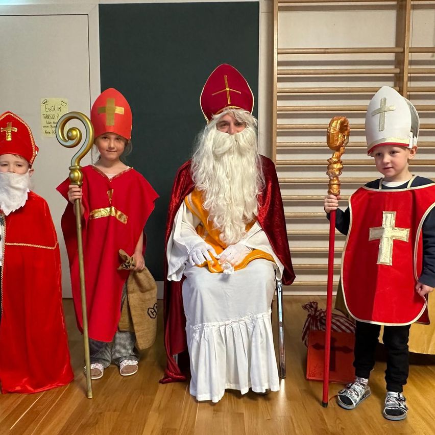 Vier Kinder in Gewändern als Heilige stehen um einen älteren Mann in einer Weihnachtsmann-Kostüm herum und halten Stäbe mit Kreuzen. Sie scheinen sich auf eine Aufführung vorzubereiten.