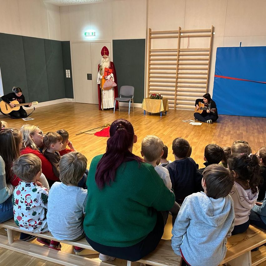 Eine Gruppe von Kindern und Erwachsenen sieht sich eine Aufführung in einem Raum an. Zwei Frauen spielen Gitarren auf der Bühne. Eine Person, die als Weihnachtsmann verkleidet ist, steht hinter einem Stuhl. Ein Tisch mit Blumen steht vor einer blauen Wand.