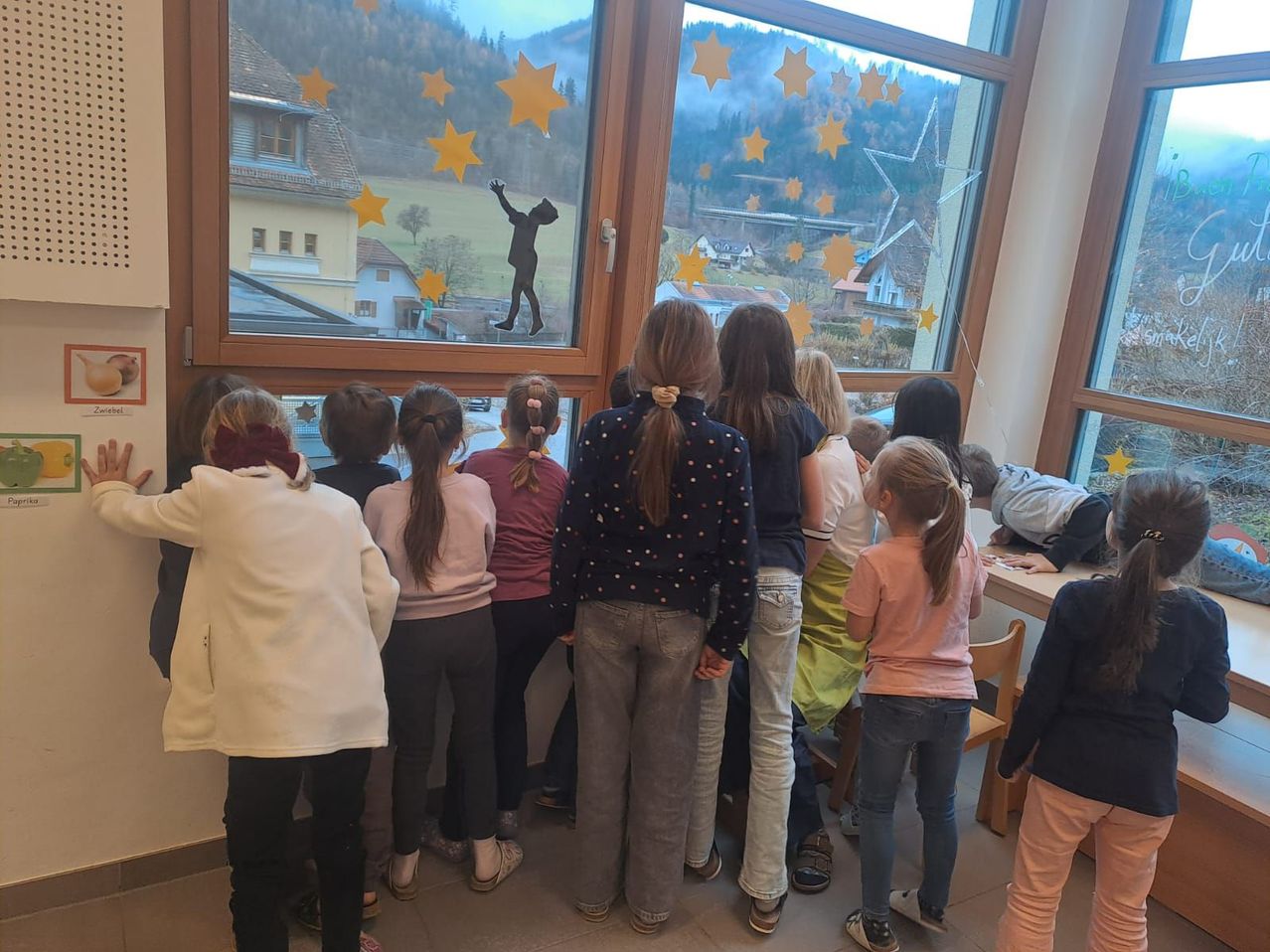 Eine Gruppe von Kindern steht vor einem Fenster und schaut nach draußen. Eine Silhouette einer Person ist mit gelben Sternen auf dem Fenster. Draußen gibt es Häuser, Bäume und Berge.
