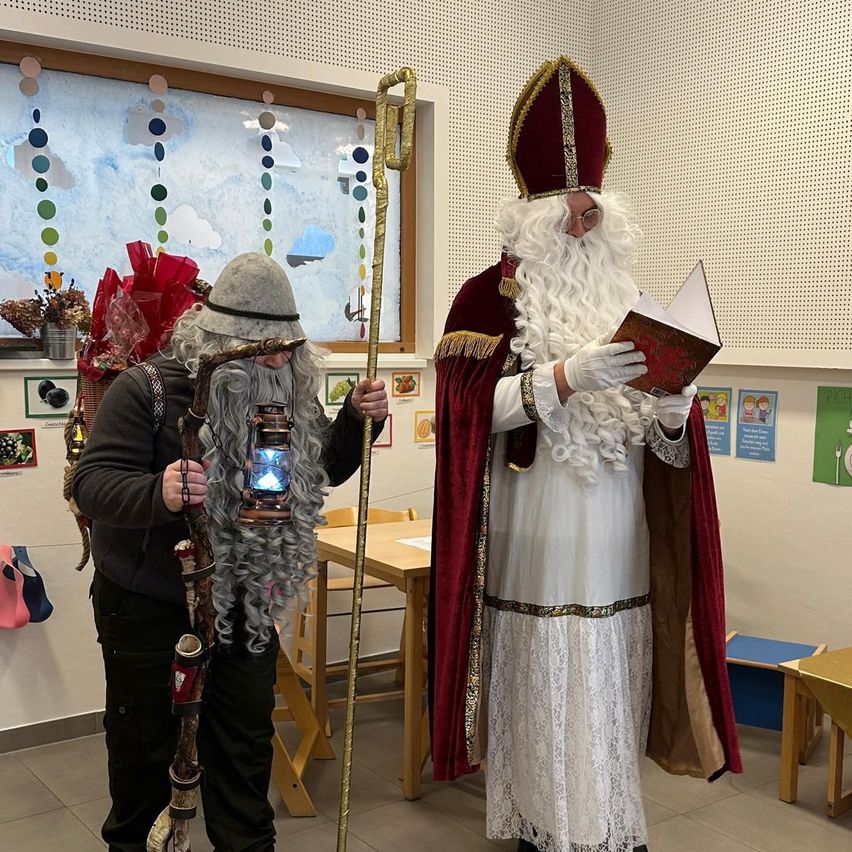 Zwei Personen in festlichen Kostümen stehen in einem Raum. Einer ist als Sankt Nikolaus gekleidet und hält ein Buch, während der andere einen Stab und eine Laterne hält. Der Raum hat Tische, Stühle und ein Fenster mit dekorativen Aufhängungen.