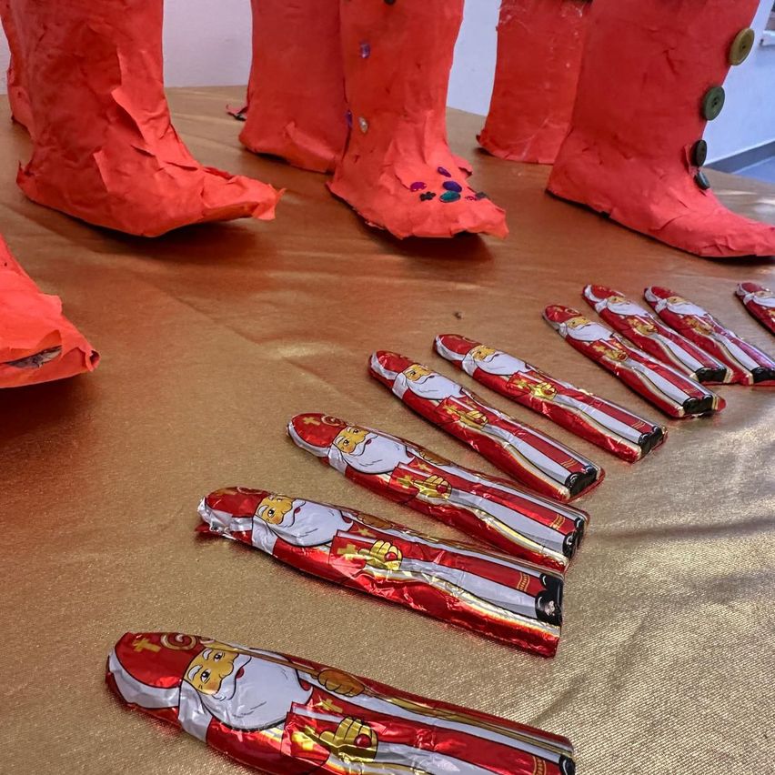 Mehrere rote Papiermaché-Stiefel und Schokoladen mit Weihnachtsmann-Designs liegen auf einer goldfarbenen Oberfläche. Die Stiefel haben bunte Knöpfe und dekorative Elemente.