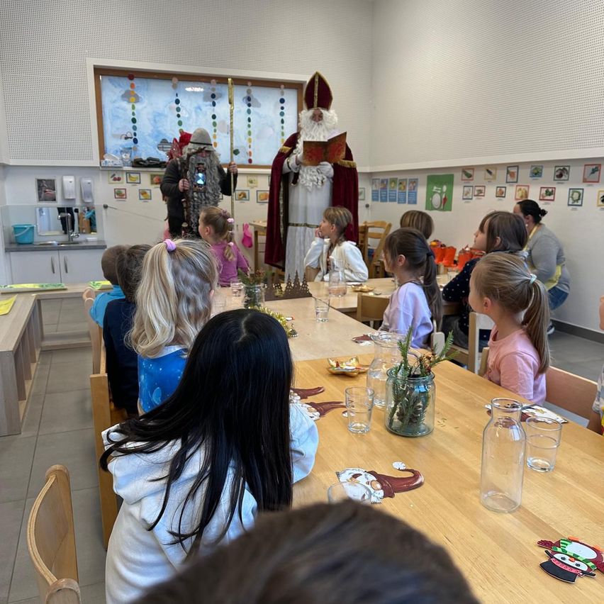 In einem Klassenzimmer sitzen Kinder an Tischen mit Gläsern, Blumen und Tellern. Ein Mann, verkleidet als Sankt Nikolaus, steht in der Nähe eines Fensters und hält ein Buch. Ein anderer Mann, verkleidet als Krampus, steht hinter den Kindern.