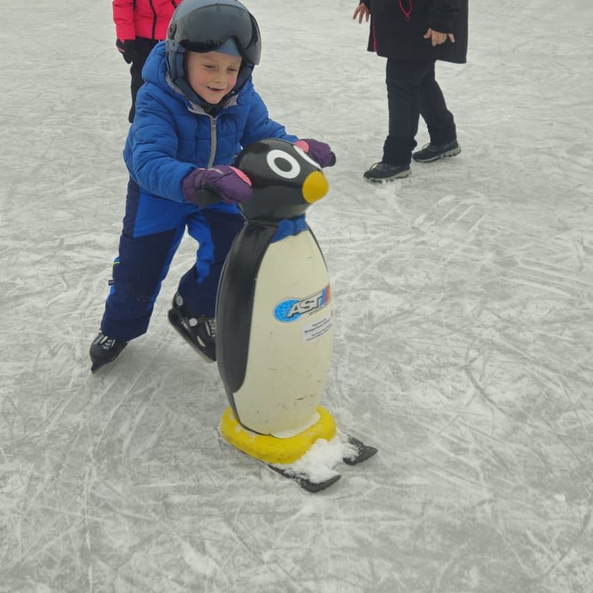 Ein junger Junge skat auf einer Eisbahn in einem Pinguinkostüm, mit zwei anderen Personen im Hintergrund.