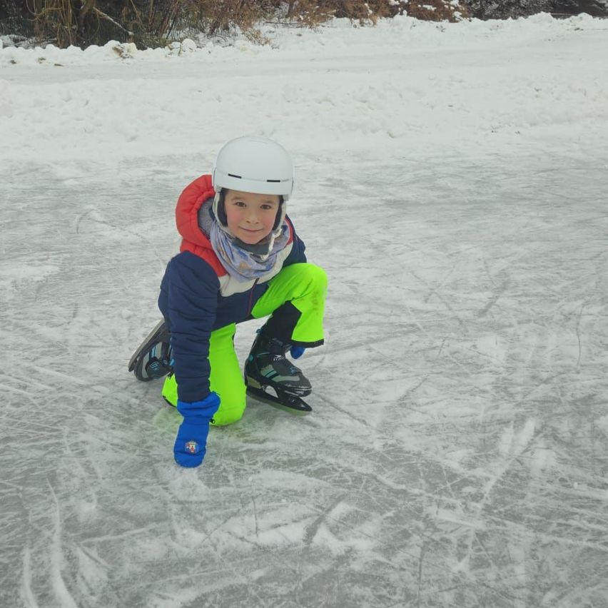 Ein junges Kind hockt auf einer Eisbahn, trägt einen Helm, Handschuhe und Schlittschuhe und lächelt in die Kamera.