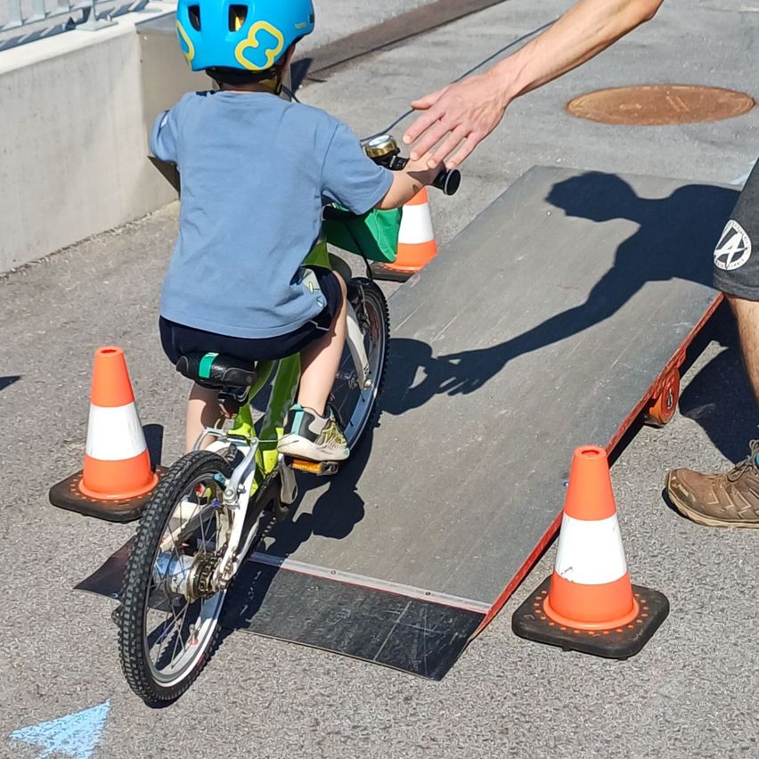 Ein Kind mit Helm fährt auf einer Rampe mit Verkehrskegel ein Fahrrad, begleitet von einem Erwachsenen.