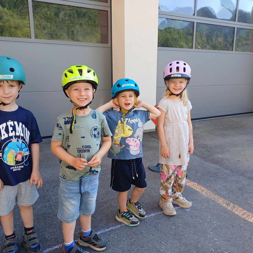 Vier Kinder stehen vor einem Gebäude und tragen alle Helme. Der Junge auf der linken Seite trägt ein blaues Shirt, der nächste Junge trägt ein grünes Shirt und der Junge daneben trägt ein hellblaues Shirt. Das Mädchen auf der rechten Seite trägt ein weißes Kleid.