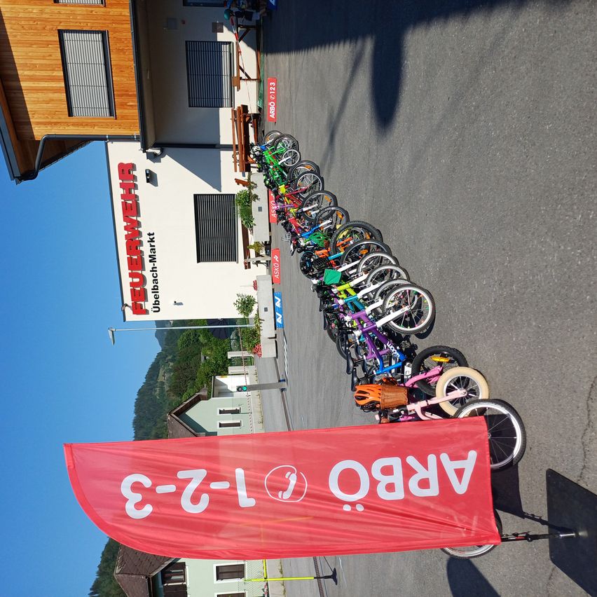 Eine Reihe bunter Fahrräder ist am Straßenrand geparkt, mit einem roten Banner, das 'E-Z-L ALBA' vor ihnen steht. Hinter den Fahrrädern ist ein Gebäude mit dem Schild 'Feuerwehr' zu sehen.