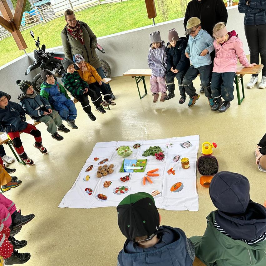 Eine Gruppe von Kindern sitzt im Kreis um einen Tisch mit verschiedenen Lebensmitteln. Ein Erwachsener steht hinter ihnen. In der Nähe ist ein Motorrad geparkt.