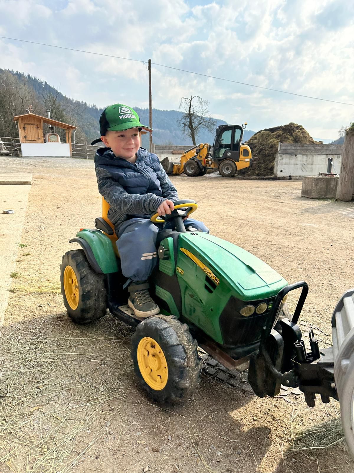Ein junger Junge mit grüner Kappe und grauem Jacken reitet auf einem grünen Traktor auf einem Feld mit einem gelben Bagger im Hintergrund.