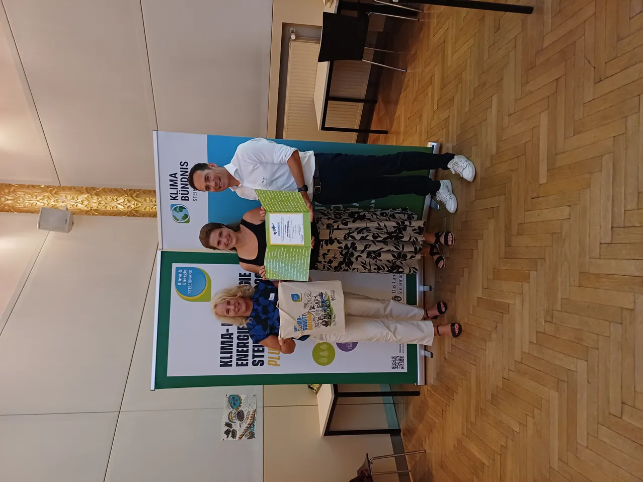 Drei Personen stehen vor einem Banner mit dem Text Klima-Bundnis und Energie-Steiermark. Die Frau in der Mitte hält ein Zertifikat und eine Tasche. Der Mann auf der linken Seite hält ein Zertifikat. Die Frau auf der rechten Seite lächelt.