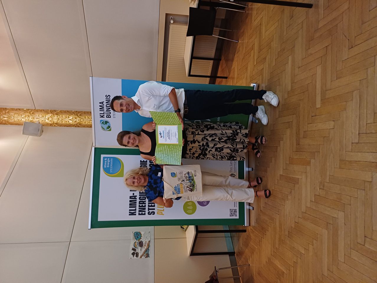Three people are standing in front of a banner with the text Klima-Bundnis and Energie-Steiermark. The woman in the middle is holding a certificate and a bag. The man on the left is holding a certificate. The woman on the right is smiling.