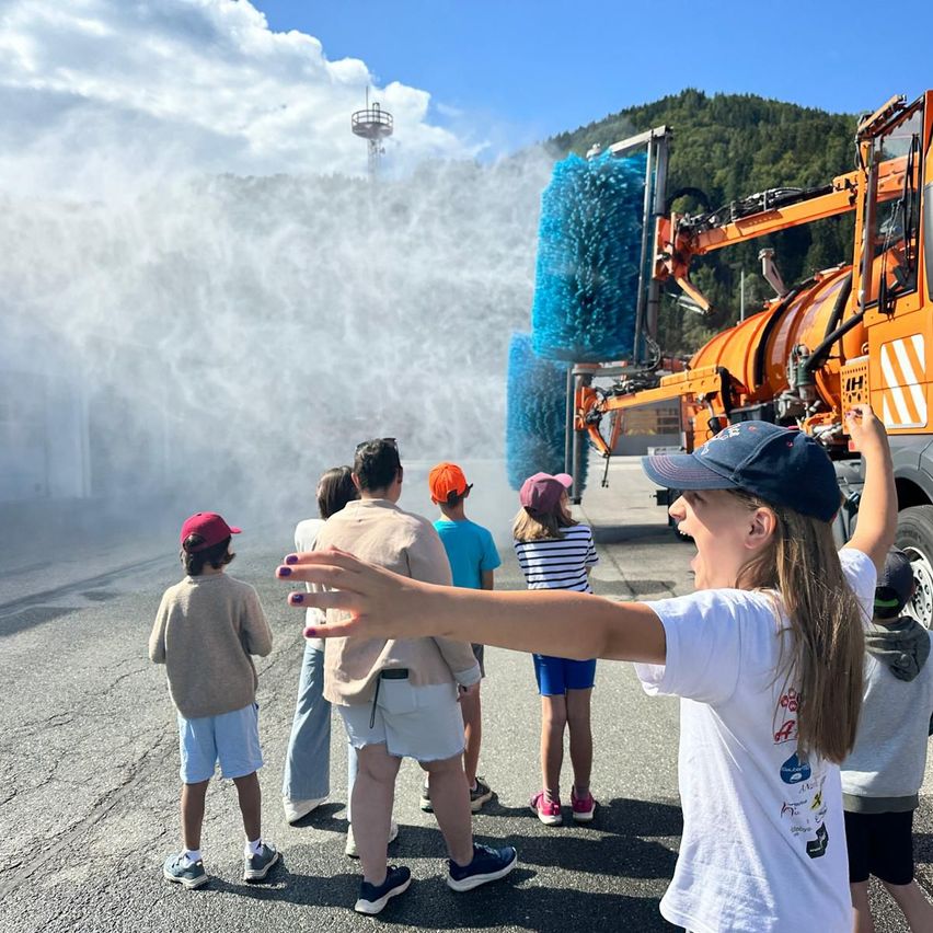 Eine Gruppe von Kindern und Erwachsenen steht vor einem großen orangefarbenen Reinigungsfahrzeug, aus dem Wasser spritzt. Die Kinder tragen Mützen und Turnschuhe, und die Erwachsenen sind lässig gekleidet. Im Hintergrund befinden sich ein Berg und ein Turm.