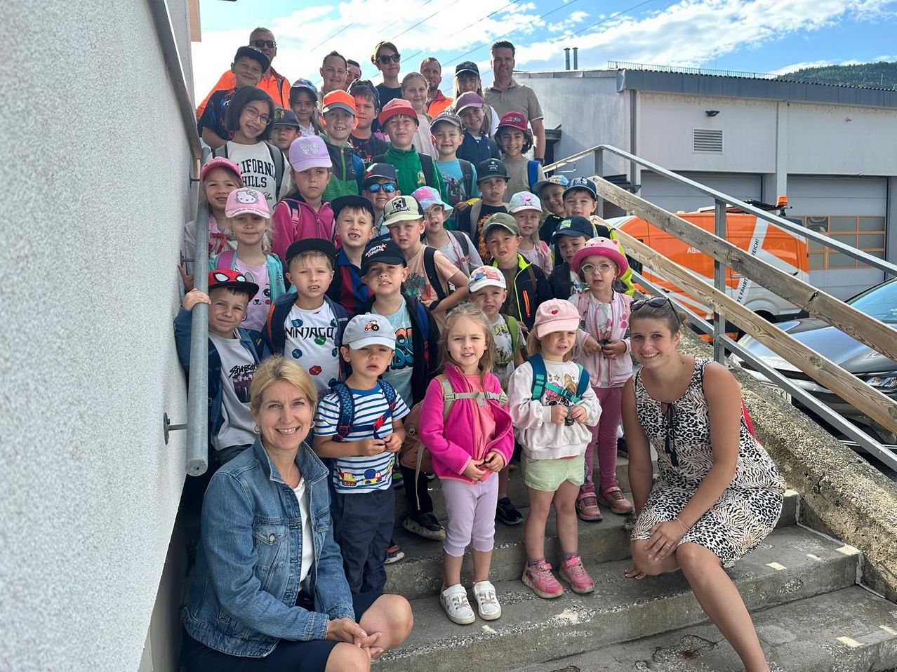 Eine Gruppe von Kindern und Erwachsenen, darunter zwei Frauen, posiert für ein Foto auf einer Treppe. Die Kinder tragen Mützen und Rucksäcke, und die Erwachsenen lächeln. Im Hintergrund sind ein Gebäude und ein Fahrzeug zu sehen.