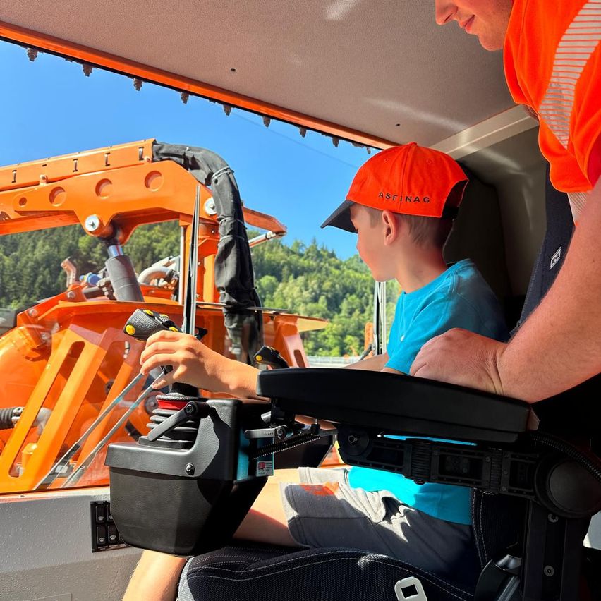 Ein junger Junge mit einer orangefarbenen Kappe sitzt auf dem Fahrersitz eines Fahrzeugs und hält das Lenkrad, während ein Erwachsener neben ihm steht.