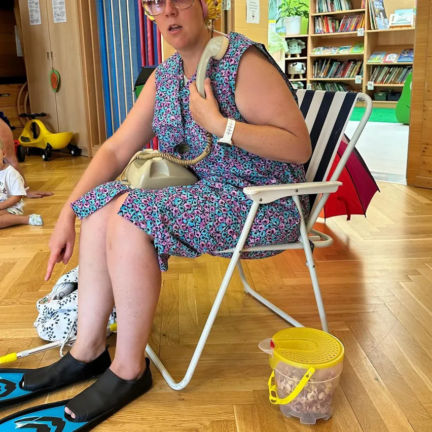 Eine Frau sitzt auf einem Klappstuhl, hält ein Telefon an ihr Ohr, trägt ein Blumenkleid und schwarze Flip-Flops. Ein Kind sitzt hinter ihr auf dem Boden.