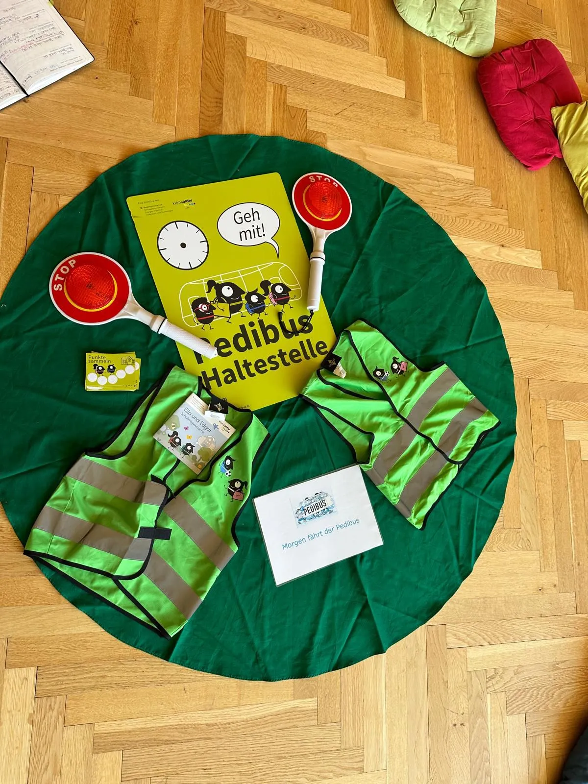 Ein grüner kreisförmiger Stoff mit Sicherheitswesten, Reflektoren und einem Buch mit dem Titel 'Geh mit!' darauf. Das Buch hat eine Uhr und eine Comicfigur.