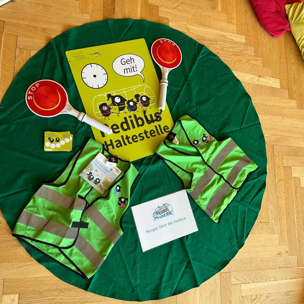 Ein grüner kreisförmiger Stoff mit Sicherheitswesten, Reflektoren und einem Buch mit dem Titel 'Geh mit!' darauf. Das Buch hat eine Uhr und eine Comicfigur.