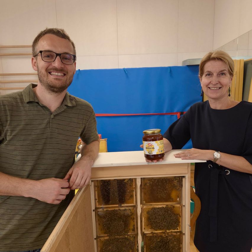 Two people stand next to a beehive inside a room, smiling. The man wears glasses and a green shirt, and the woman wears a black dress and a watch. A jar of honey sits on the beehive. Behind them is a blue wall with a blue curtain.