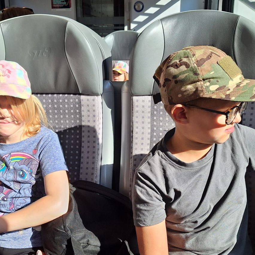 Zwei Kinder sitzen in einem Bus, ein Mädchen mit einer Regenbogenmütze und ein Junge mit einer Tarnmütze. Sie sitzen sich gegenüber mit einem Sitz dazwischen.