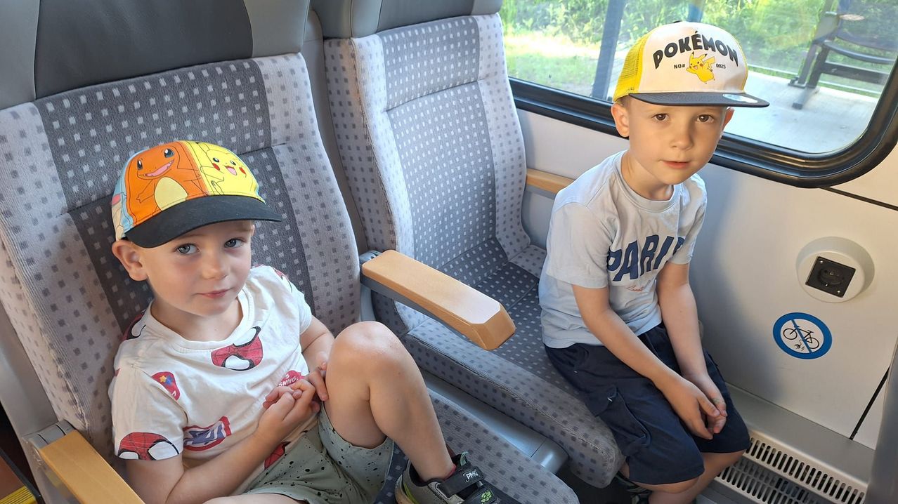 Zwei Jungen sitzen in einem Zug, einer trägt eine Pikachu-Mütze, der andere ein Shirt mit 'PARI'. Beide schauen in die Kamera, das Fenster zeigt ein Grasfeld.