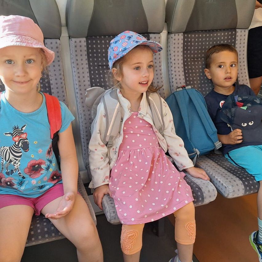 Drei junge Kinder, die Mützen und Rucksäcke tragen, sitzen zusammen in einem Bus. Das Mädchen in der Mitte trägt ein rosa gepunktetes Kleid. Der Junge rechts trägt ein blaues Hemd und hält eine Mütze.