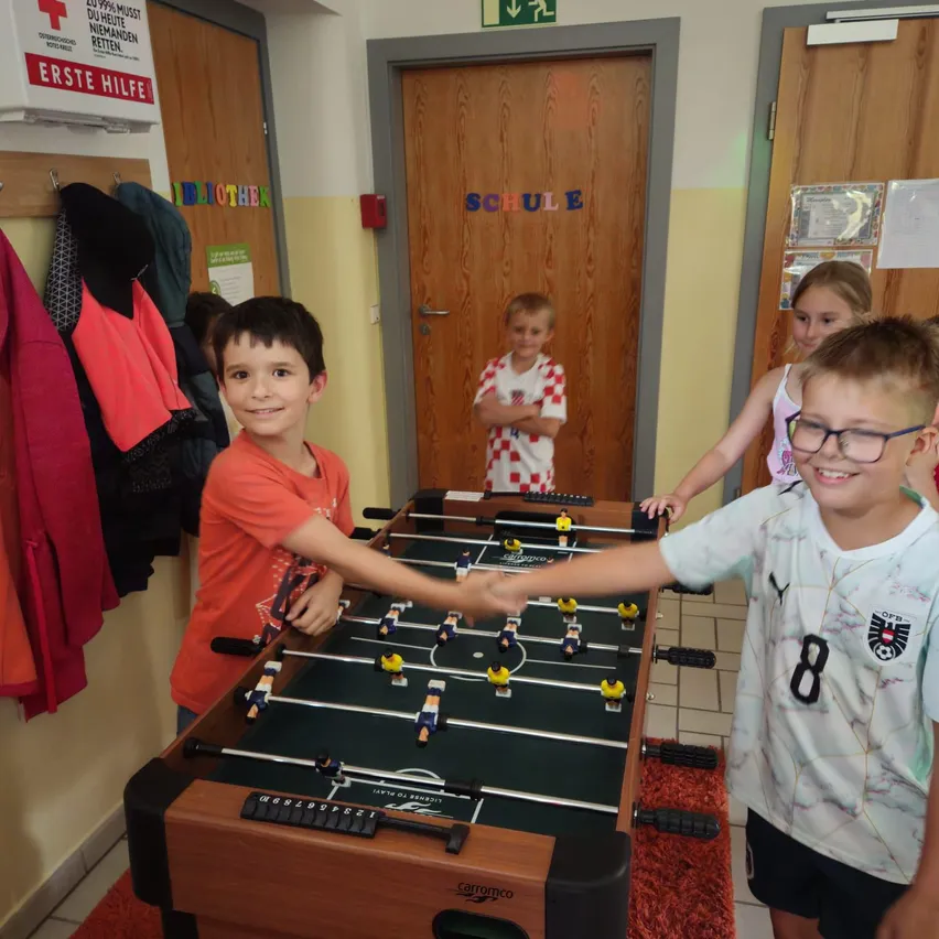 Vier Kinder spielen Tischfußball in einem Raum mit Holztüren und gelber Wand. Der Junge im orangefarbenen Shirt schüttelt dem Jungen im weißen Shirt die Hand.