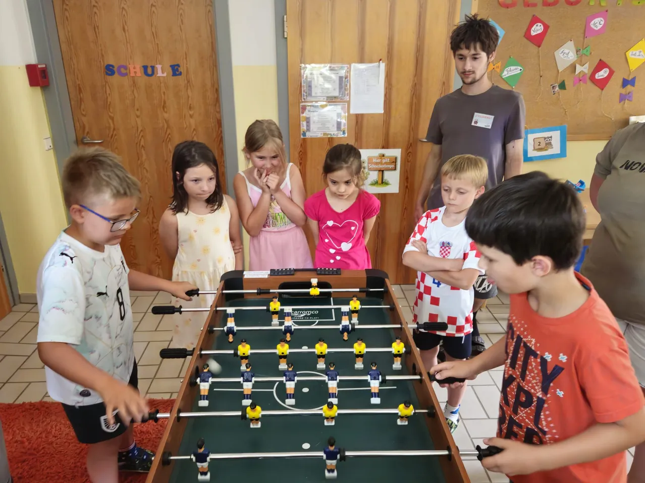 Eine Gruppe von Kindern spielt Tischfußball in einem Klassenzimmer, wobei ein Lehrer beobachtet. Die Kinder sind engagiert und konzentrieren sich auf das Spiel.