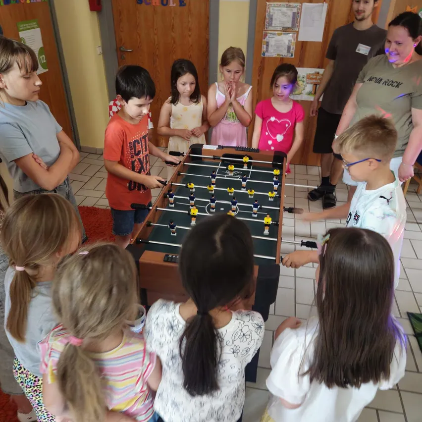 Eine Gruppe von Kindern und Erwachsenen versammelt sich um einen Tischkicker in einem Raum. Einige schauen zu, während andere spielen.