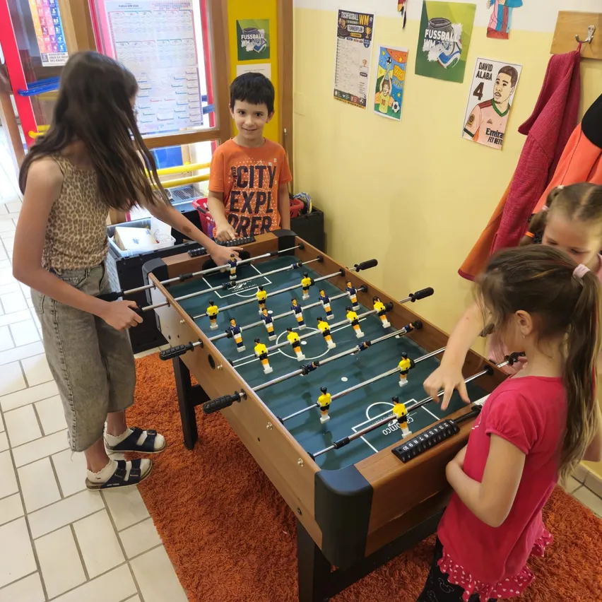 Vier Kinder spielen Tischfußball in einem Raum mit gelber Wand, Teppich und Postern an der Wand.